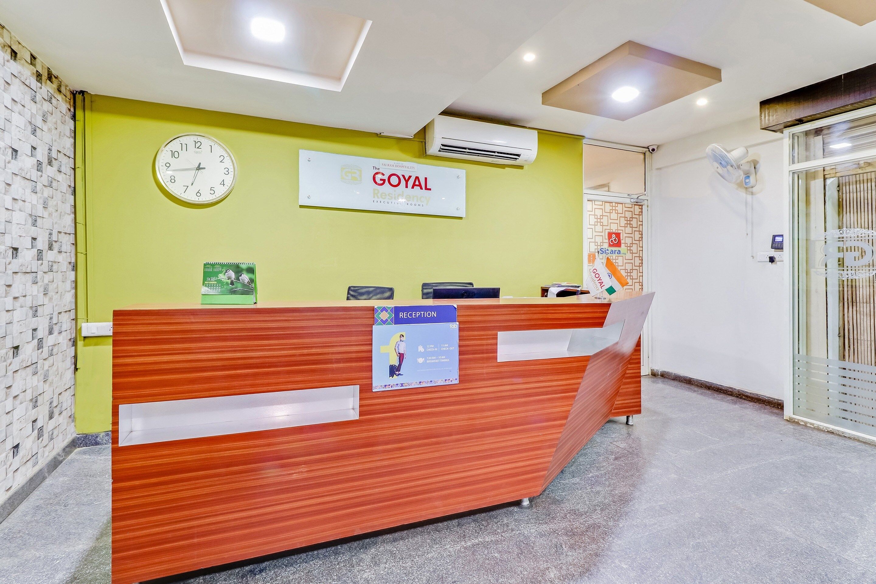 Un mostrador de recepción con un reloj, un letrero que dice 'The Goyal Residency Executive Rooms' y una zona de recepción con puerta de vidrio.