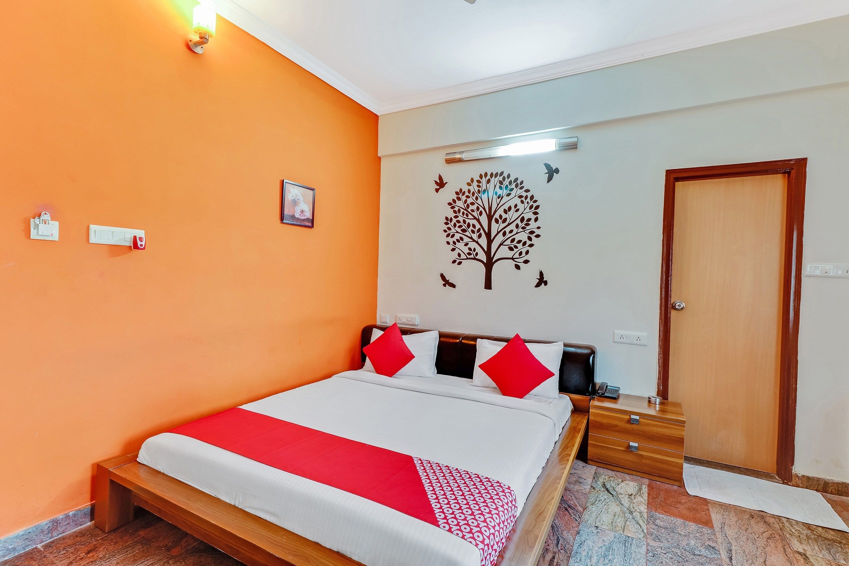 Habitación de hotel con pared naranja, cama con ropa de cama blanca y roja, cabecero de madera, luz fijada a la pared, puerta y una mesita con un teléfono.