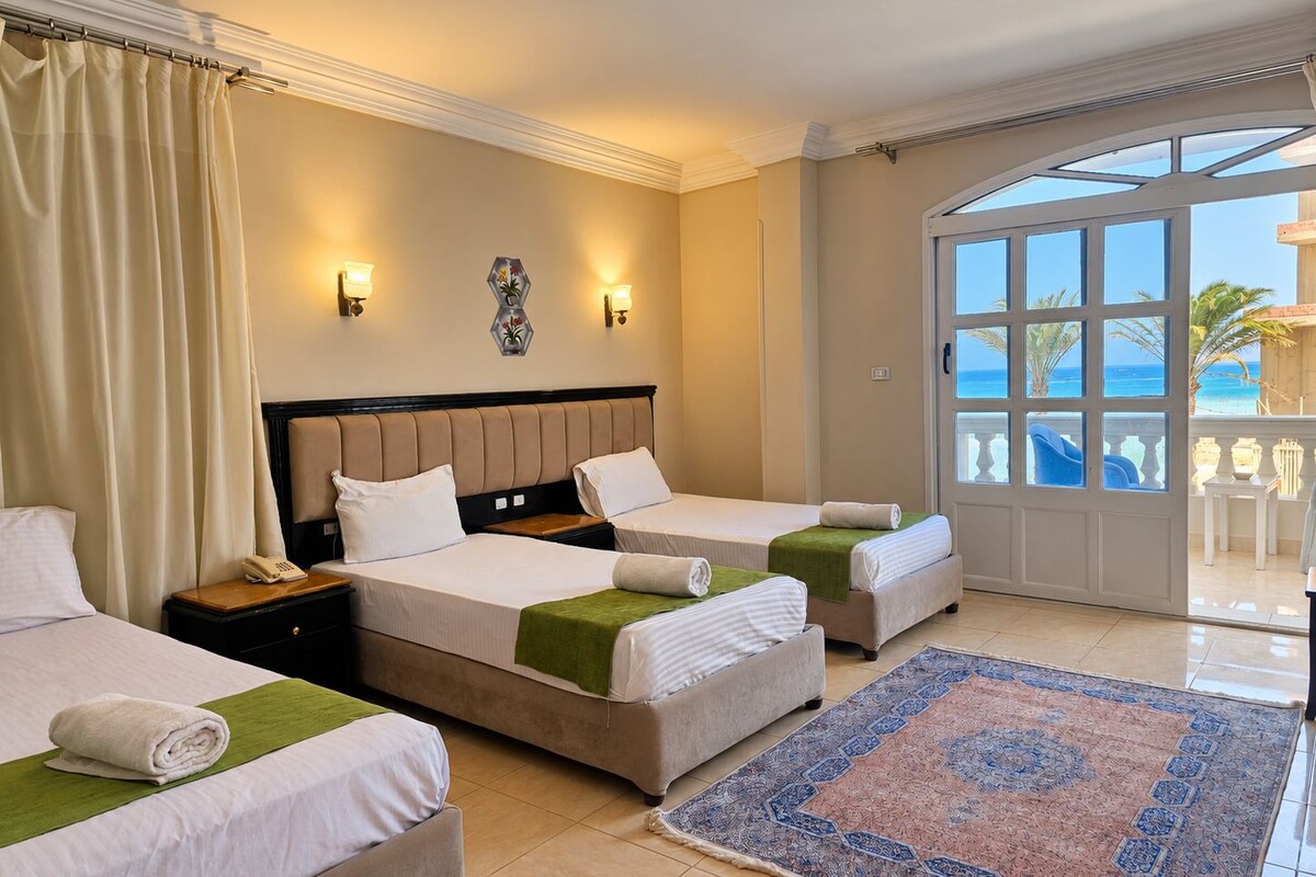 A hotel room with two beds, a large window with a view of the sea, and a balcony with outdoor furniture.