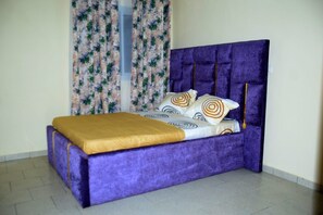 Una cama con cabecera tapizada en morado, colcha estampada y detalles dorados.
