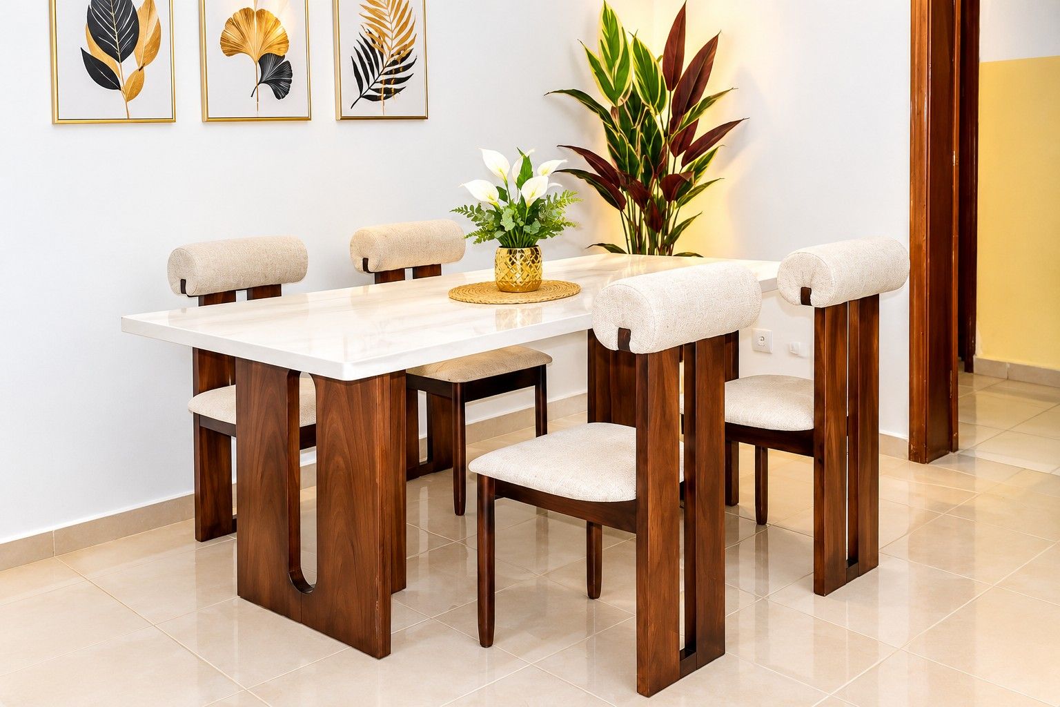 A dining area with a white table and wooden chairs, a vase with flowers, and framed leaf art on the wall.