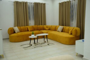 Una sala de estar con un sofá color mostaza, dos mesitas redondas, un armario blanco y cortinas color beige.