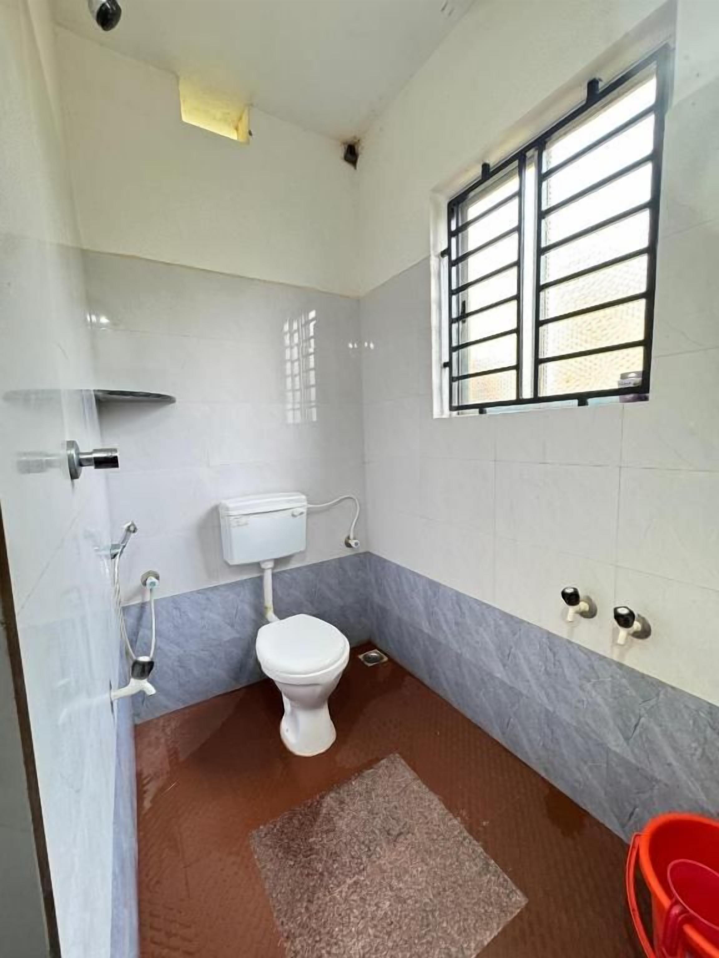 Un baño con inodoro, una ventana con barrotes, un cubo rojo y zona de ducha.