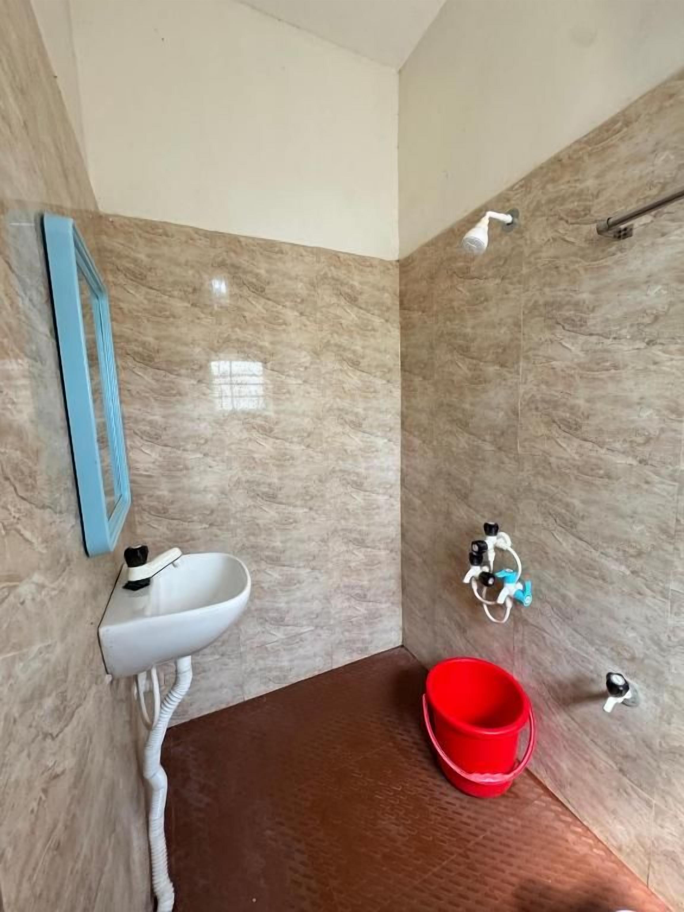 Un baño con pared de mármol, un lavamanos blanco, un cubo rojo y un cabezal de ducha.