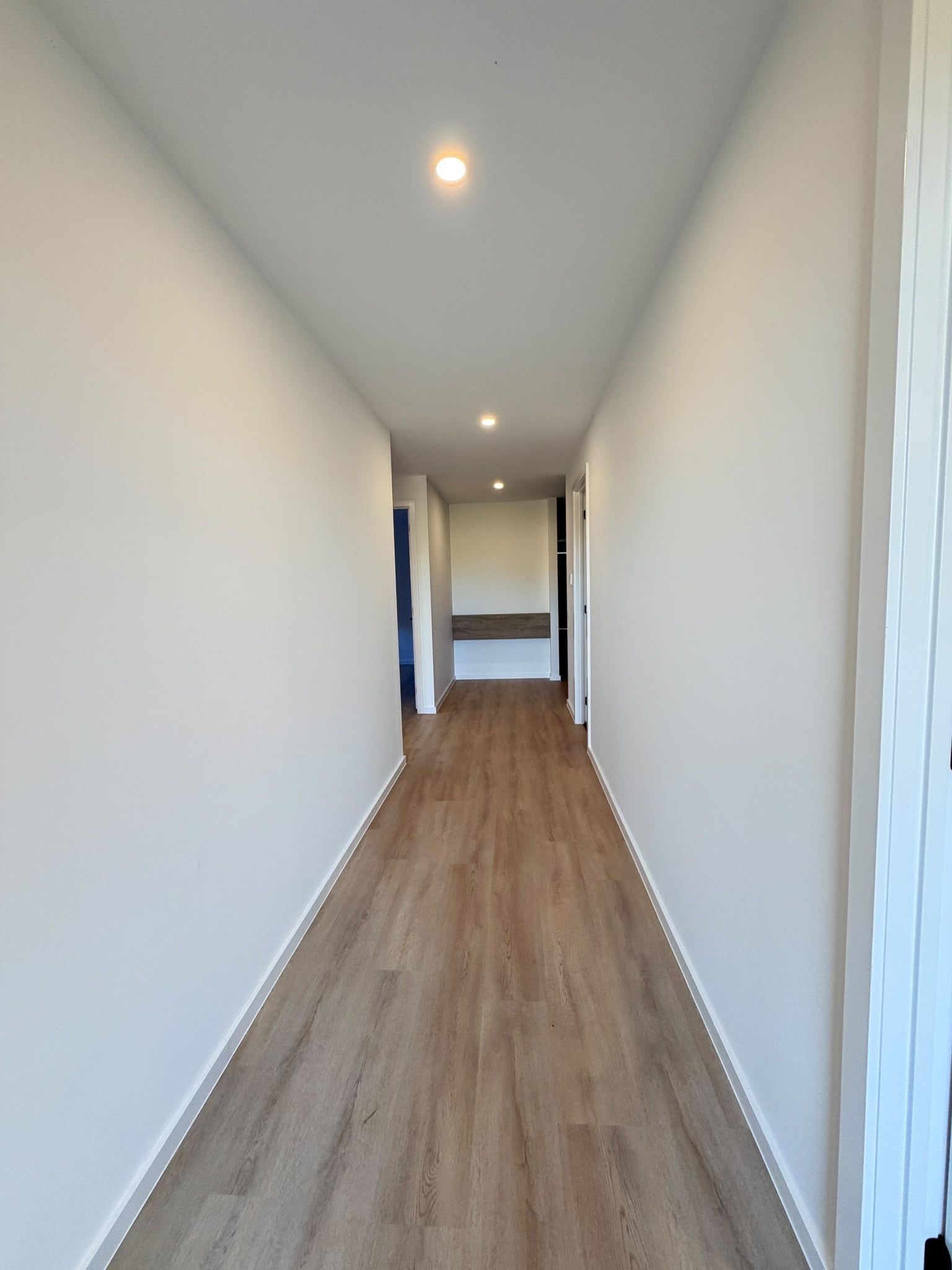 A hallway with wooden flooring and white walls, leading to a room with a door at the end.
