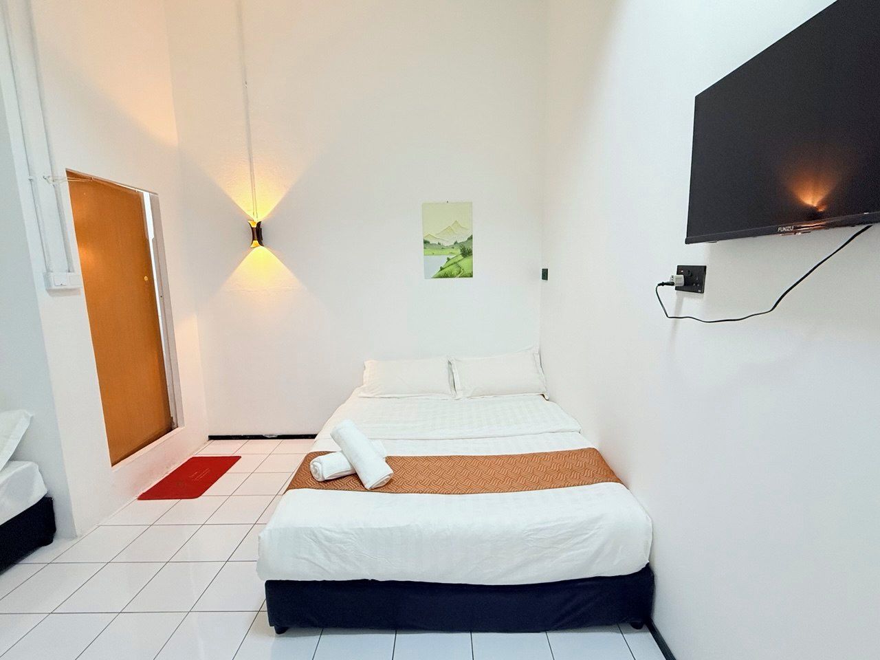 Habitación de hotel con cama, televisor de pantalla plana en la pared, una puerta y un pequeño cuadro colgado.