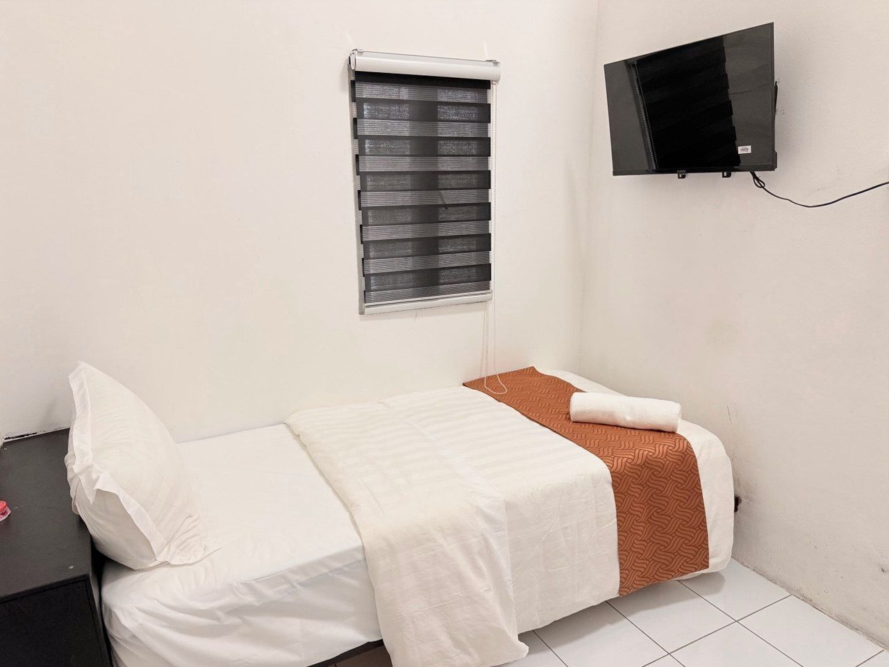 Ein kleines Zimmer mit einem Einzelbett, einem Fenster, einem an der Wand befestigten Flachbildfernseher und einem Nachttisch mit Lampe.