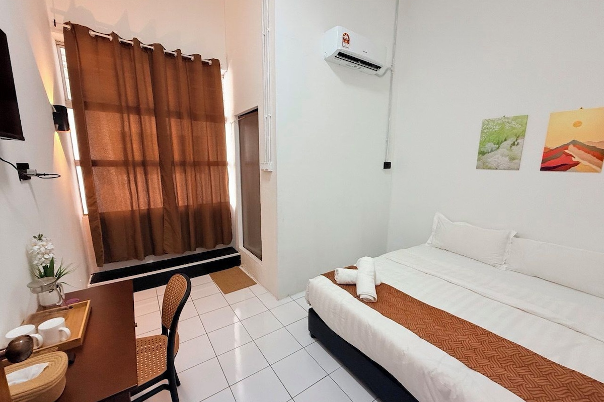 A hotel room with a bed, a desk with a chair, a window with curtains, and wall-mounted air conditioning.