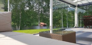 A modern outdoor seating area with a glass enclosure, a wicker table, and a view of a natural landscape. - River landscape close to the centre, Himos 8km (Jämsä)