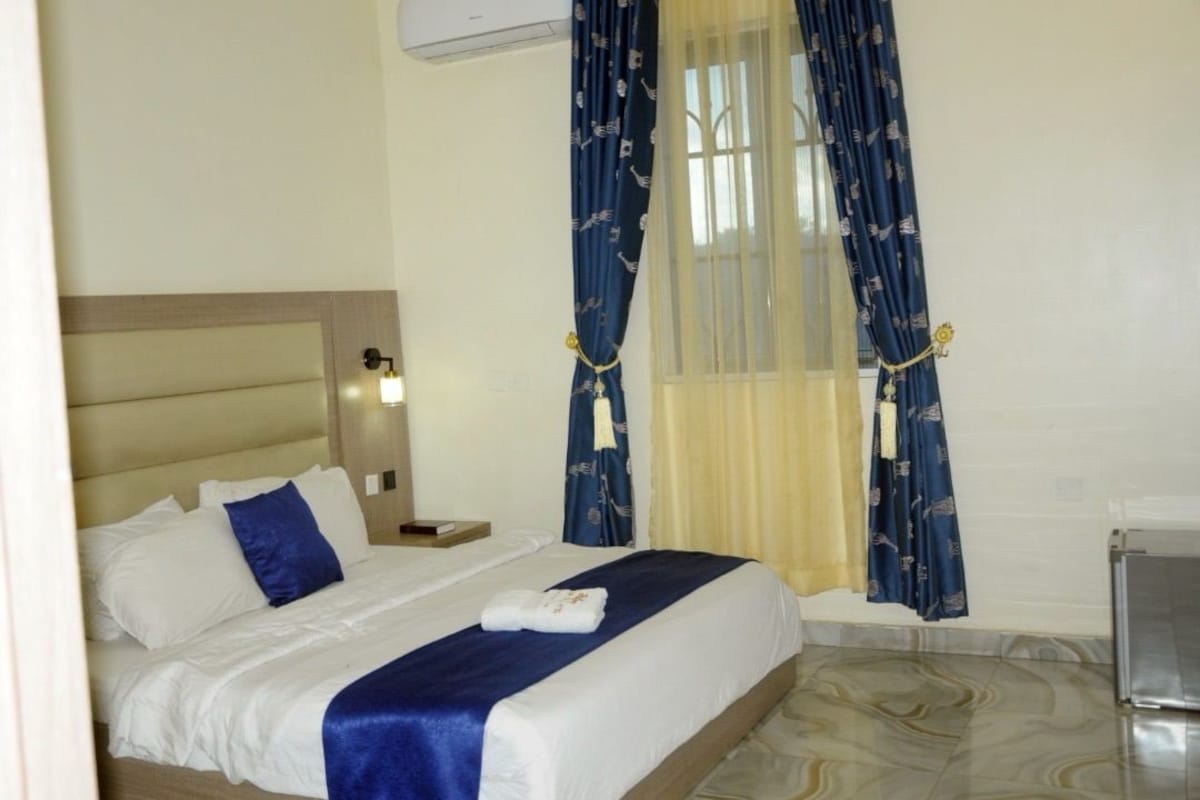 Quarto de hotel com cama, mesa de cabeceira, luminária, janela com cortinas e um espaço para o banheiro.