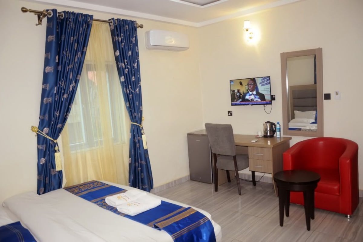Quarto de hotel com cama, escrivaninha, cadeira, poltrona vermelha e televisão.