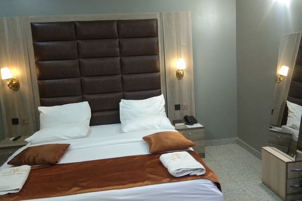 Quarto de hotel com uma cama grande, duas mesas de cabeceira, um criado-mudo, um espelho e luminárias fixadas na parede.