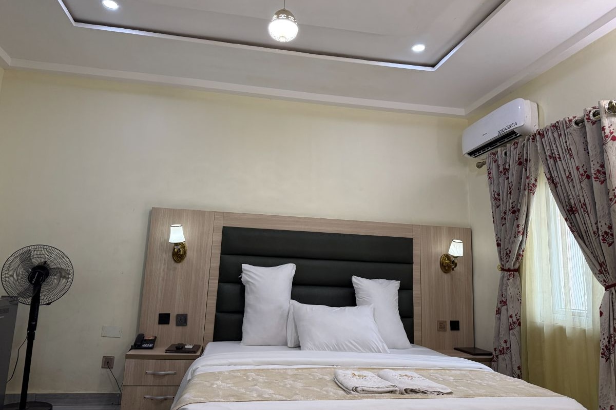 Quarto de hotel com cama, abajures, ventoinha, ar condicionado e janela com cortinas.