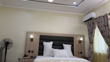 A hotel room with a bed, bedside lamps, a fan, an air conditioning unit, and a window with curtains.