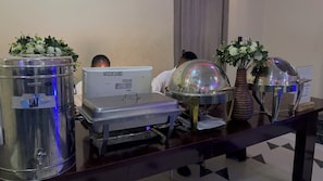 Disposição de buffet com recipientes de aço inoxidável, um dispensador de água e um vaso decorativo com flores.
