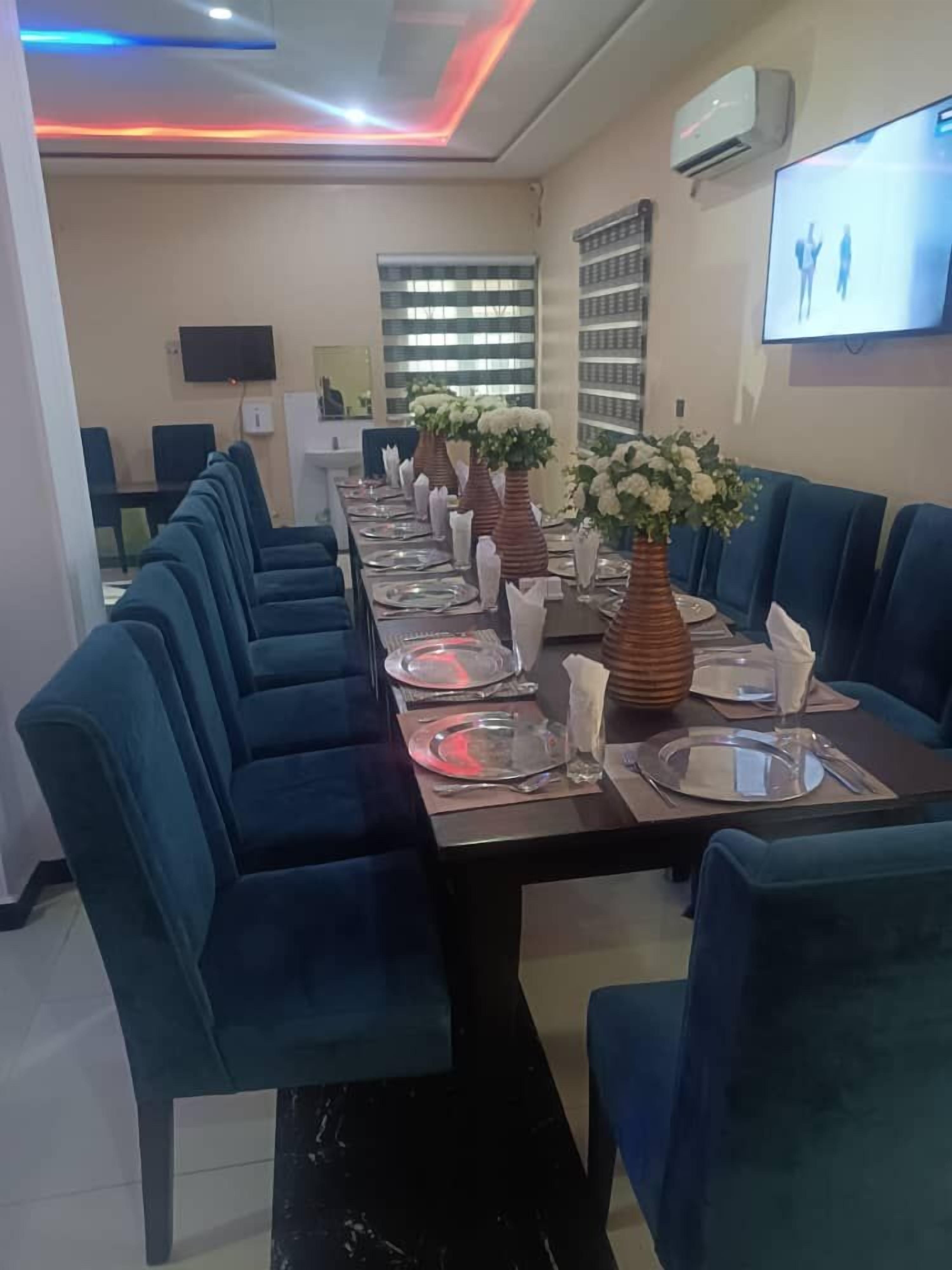Una larga mesa de comedor con sillas azules, un televisor de pantalla plana montado en la pared y una ventana con persianas.