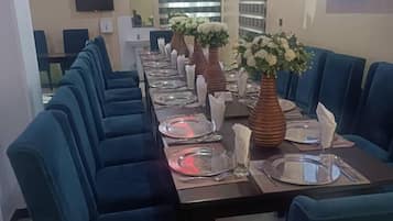 Una larga mesa de comedor con sillas azules, un televisor de pantalla plana montado en la pared y una ventana con persianas.