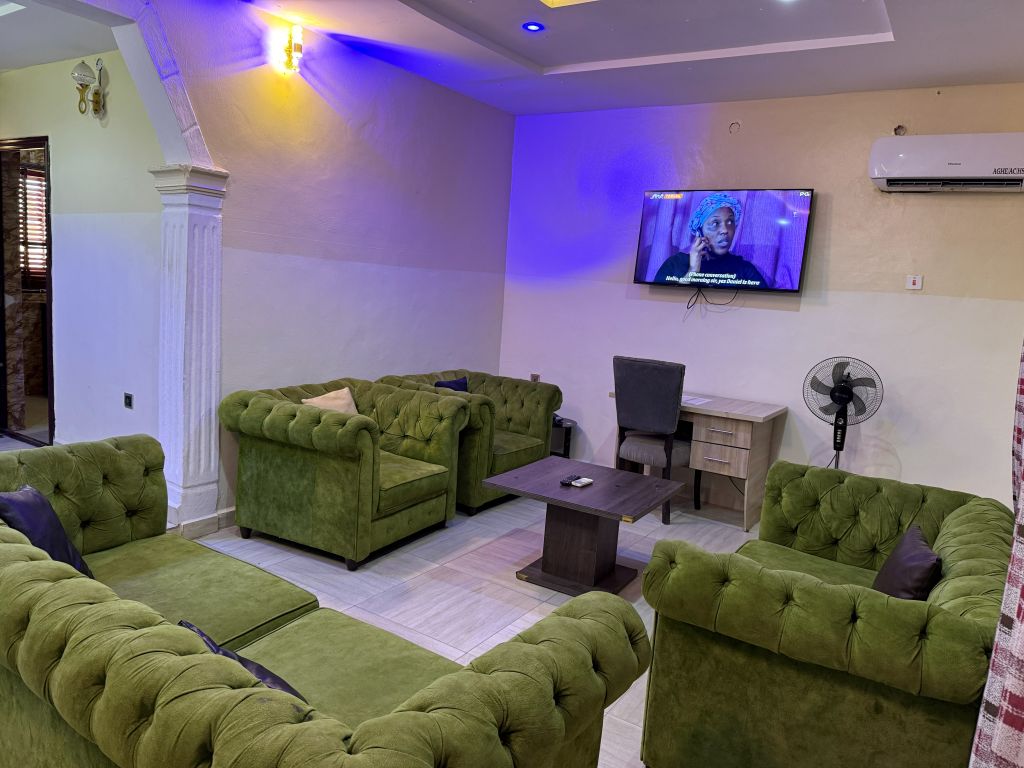 Una sala de estar con sofás tapizados en verde, un televisor de pantalla plana montado en la pared, un escritorio con una silla y un ventilador de pie.