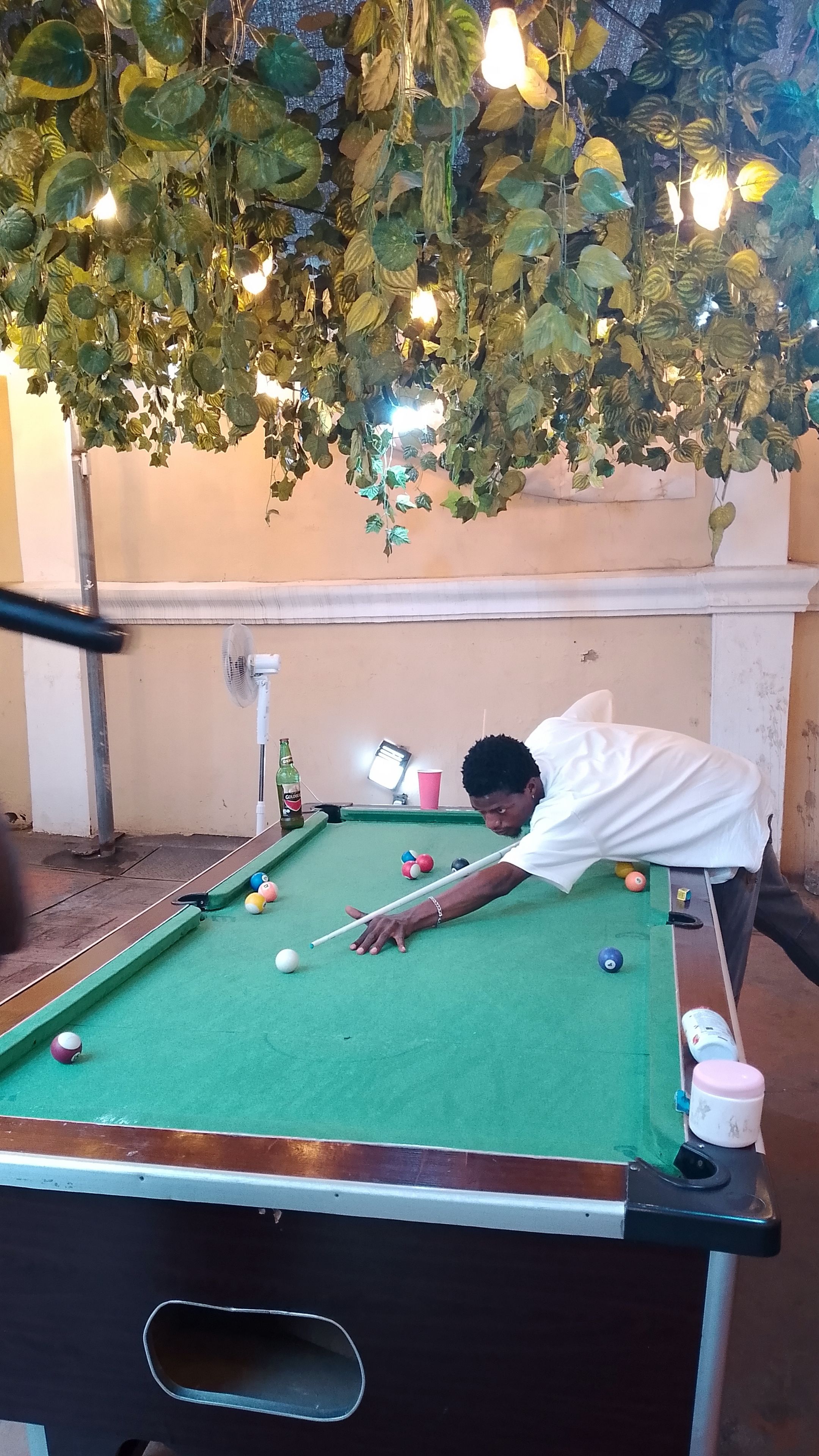 A pool table with a person playing, a fan, and a bottle of beer.