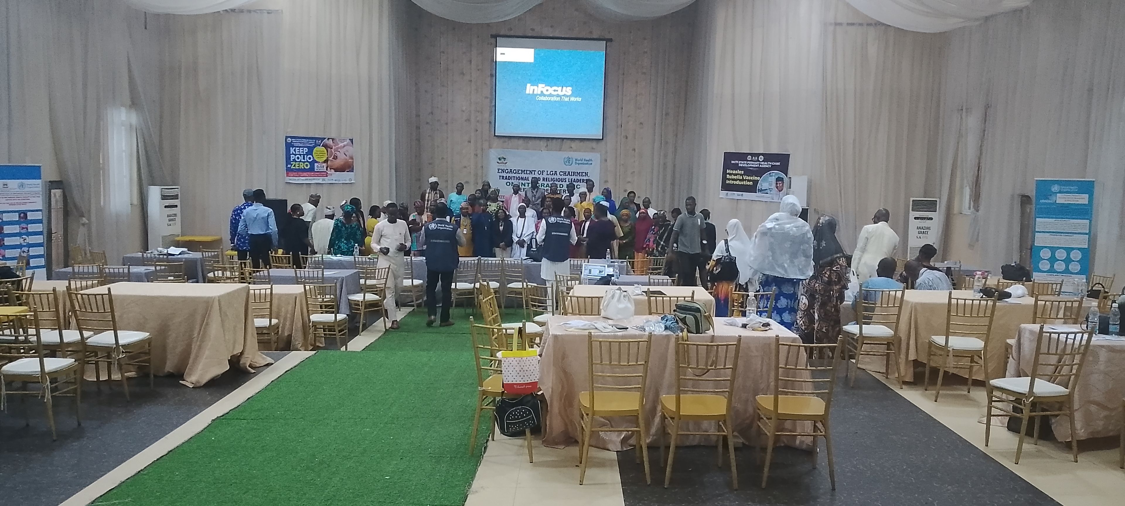 A conference room with rows of tables and chairs, a stage with a screen, and banners on the walls.