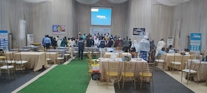 Un salón de conferencias con filas de mesas y sillas, un escenario con una pantalla y carteles en las paredes.
