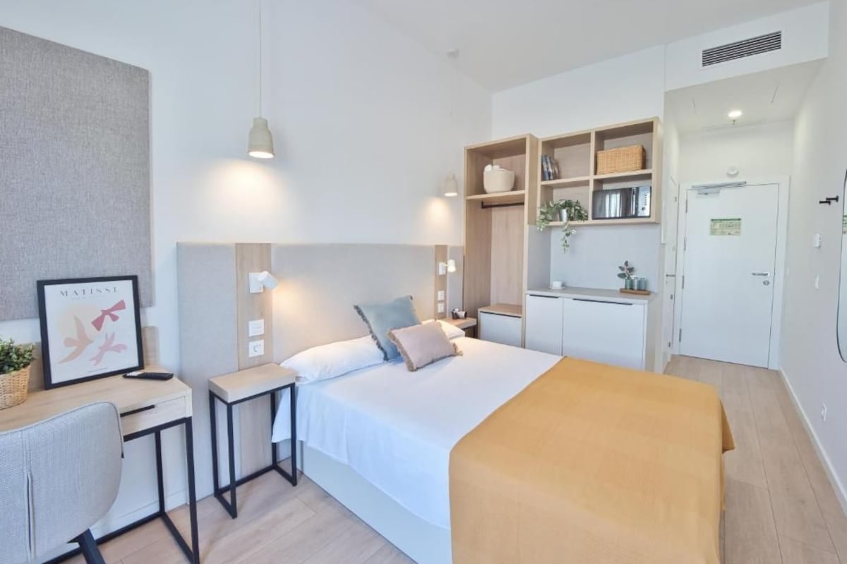A modern hotel room with a bed, desk, and kitchenette.