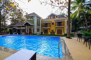 A swimming pool with a clear blue water surface, surrounded by a two-story building with balconies, and a patio area with outdoor seating. - The Amrit Goa (Candolim)