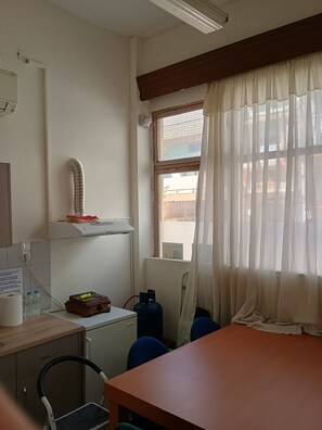 A room with a table, chairs, a heater, a window with curtains, and a wall-mounted shelf. - thomasbnb alexandroupolis (Alexandroupoli)
