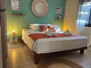 A bed with white bedding, a red throw blanket, and rolled towels. There is a small stool with a candle, a mirror with a decorative frame, and a shelf with a lantern. - New Listing (Saint-Paul)