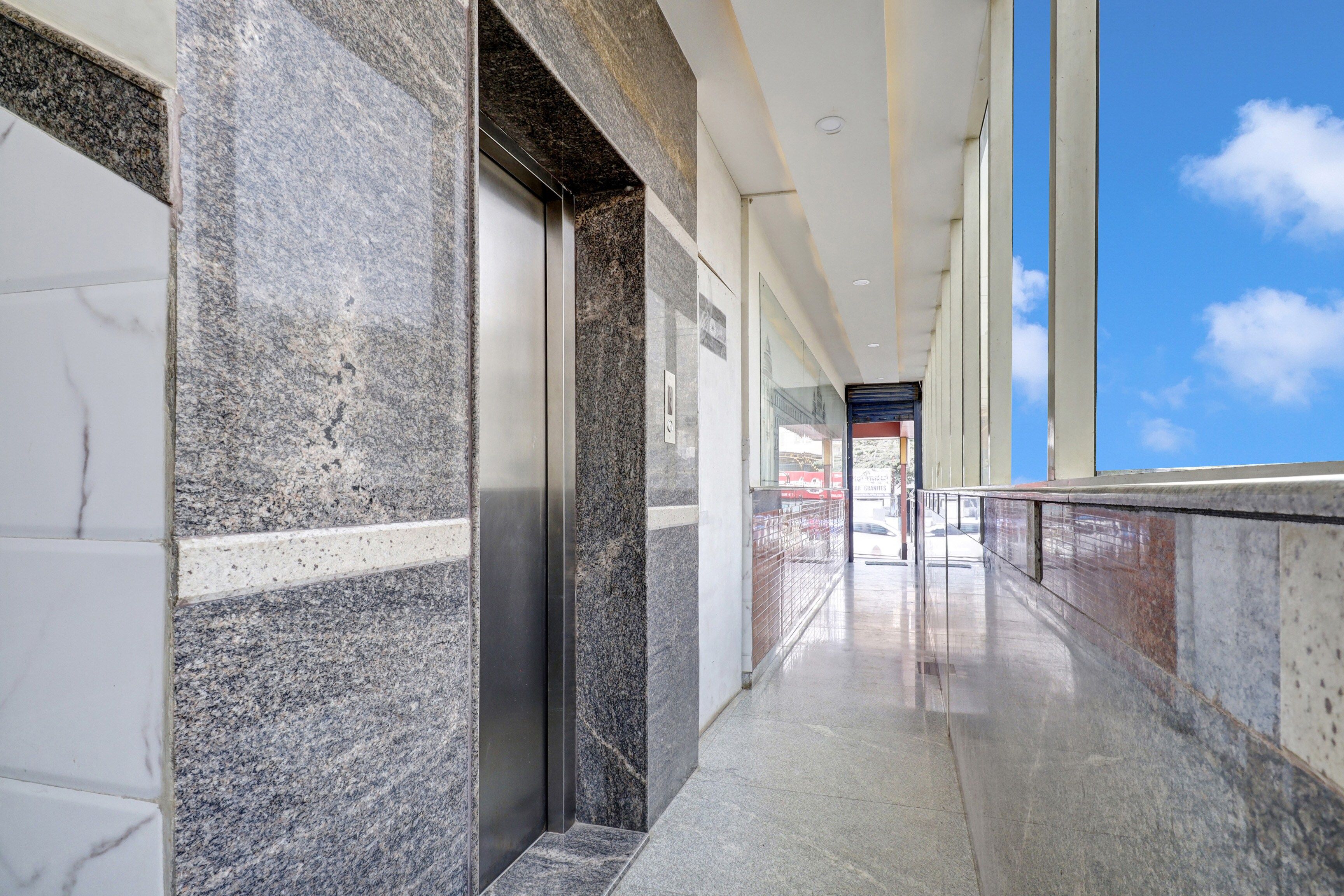 A corridor with a marble wall, an elevator, and a glass door leading to an outdoor area.