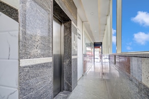 Un pasillo con pared de mármol, un ascensor y una puerta de cristal que da a un espacio exterior.