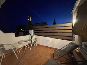 Un espace extérieur aménagé en terrasse, avec une table et des chaises, un pot de fleurs et une vue sur un bâtiment doté d’un clocher.