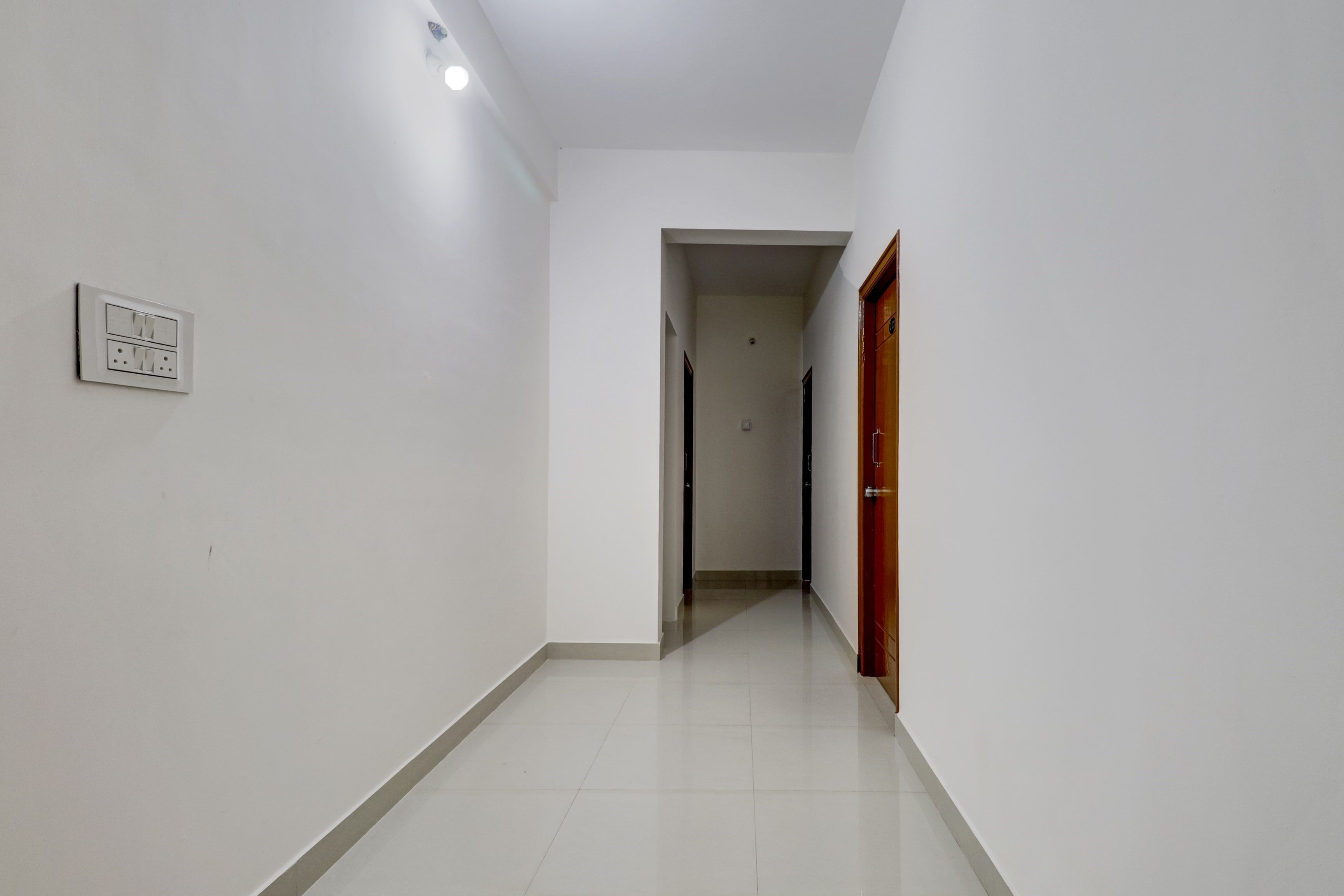 A hallway with a light switch on the wall, a door at the end, and a tiled floor.