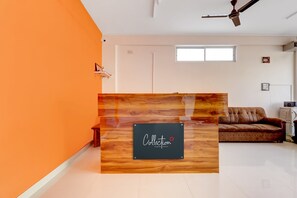 Un comptoir d’accueil avec un panneau indicateur, un ventilateur de plafond, une fenêtre et un canapé dans une pièce dont un mur est peint dans une teinte orange.
