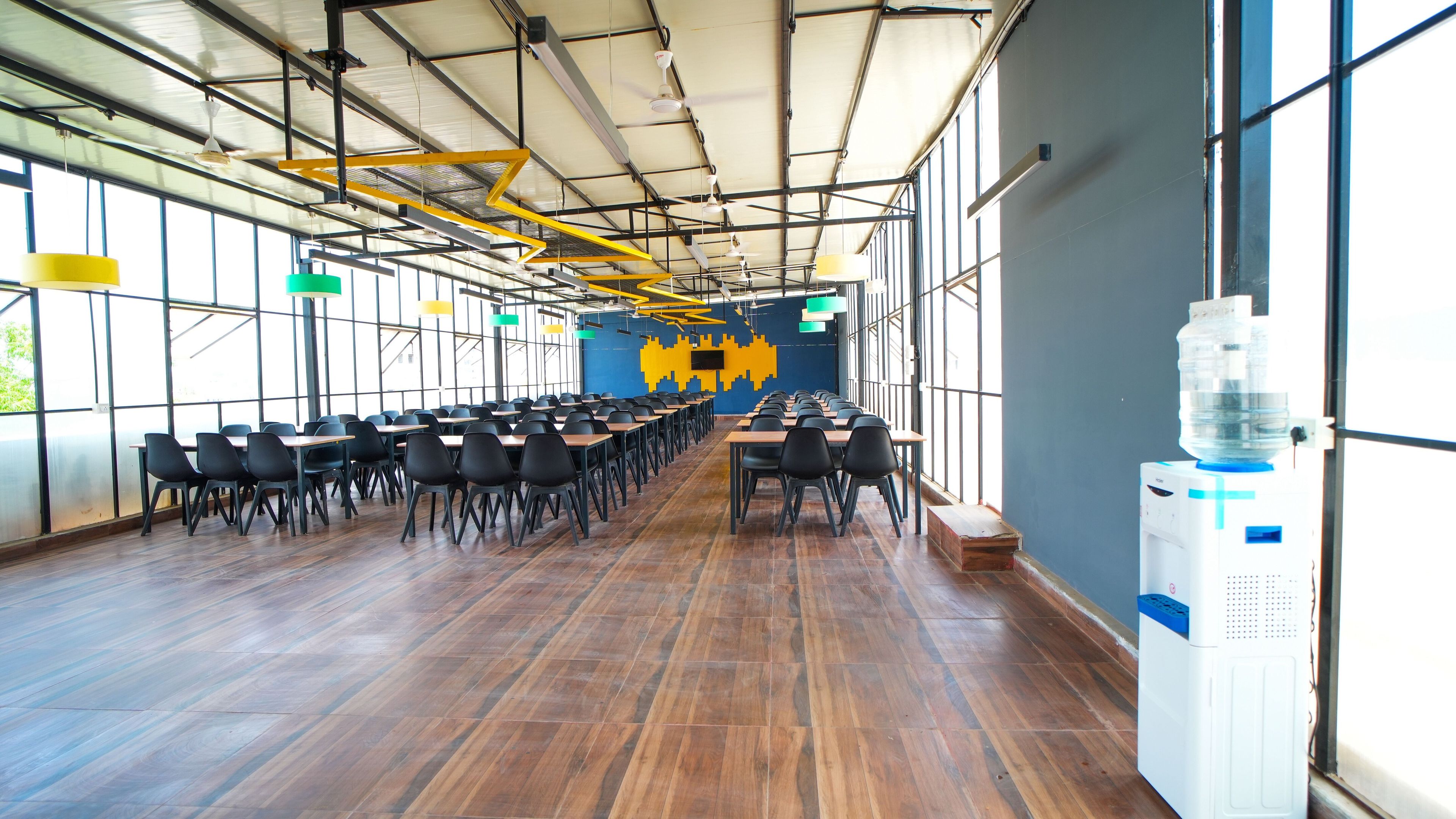 A spacious indoor area with rows of tables and chairs, large windows, and a ceiling with exposed beams and lighting fixtures.