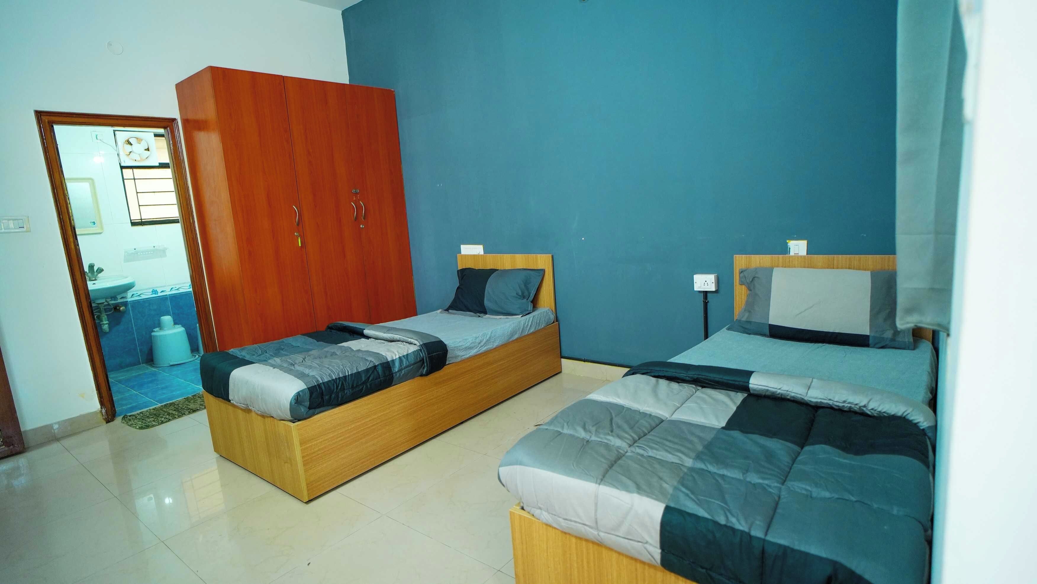 A bedroom with two single beds, a wooden wardrobe, a bathroom visible through an open door, and a tiled floor.