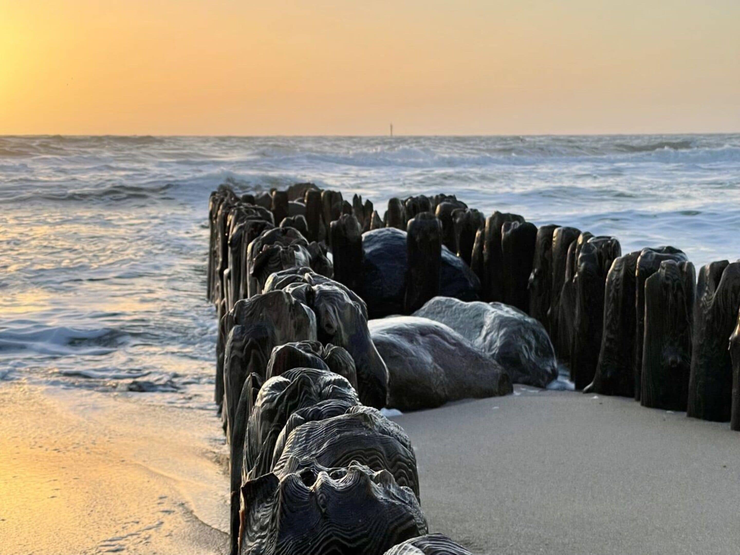 Un paysage côtier avec une digue en bois qui s’avance dans la mer, une plage de sable et les vagues de l’océan.
