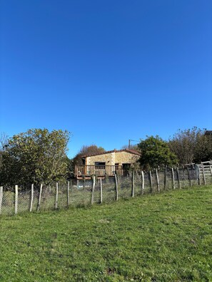 Un bâtiment en pierre doté d’une terrasse en bois, entouré d’une clôture en treillis métallique et de verdure.