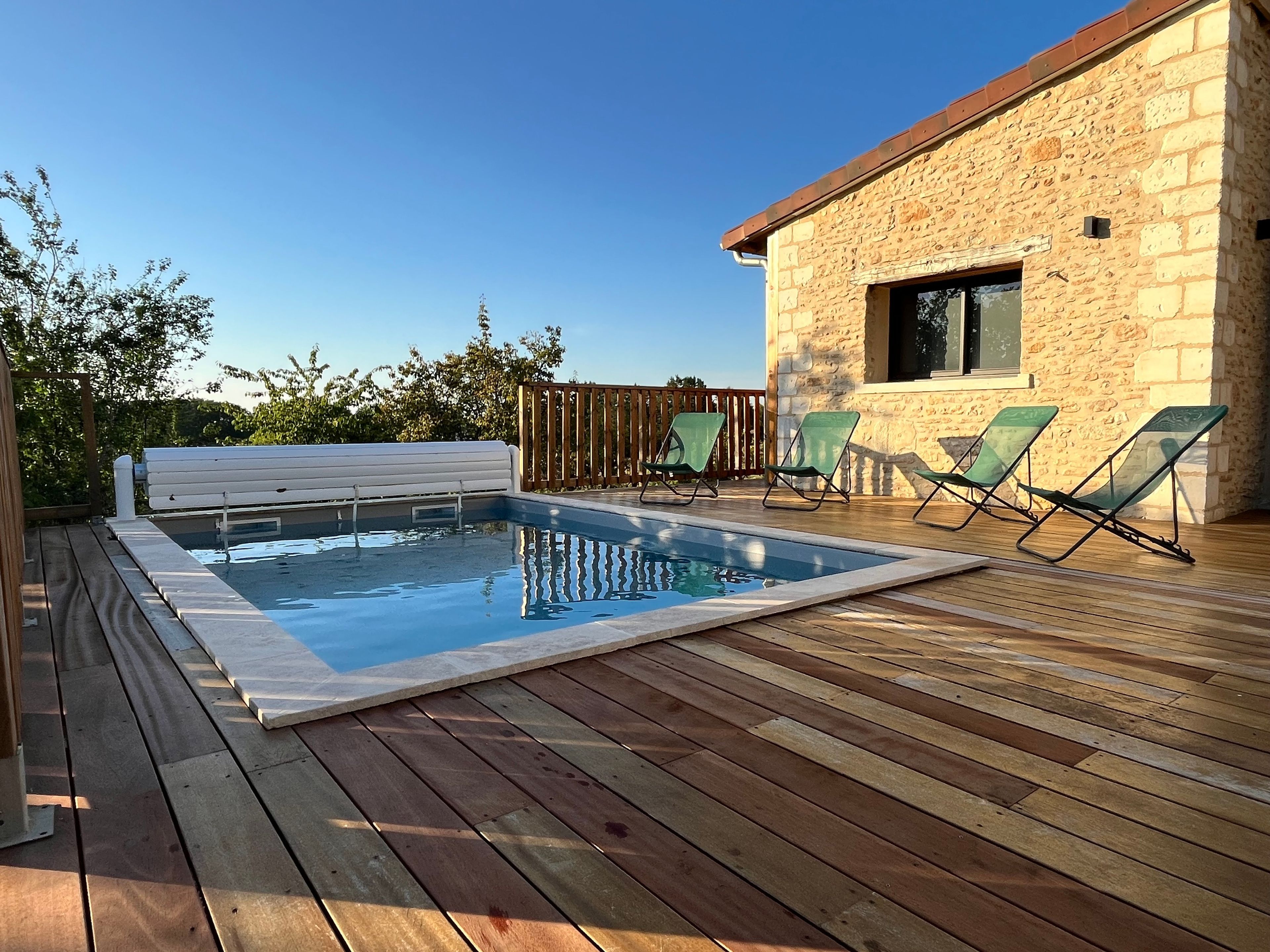 Ein Schwimmbecken mit Holzterrasse, ein Steinhaus und zwei grüne Liegestühle.