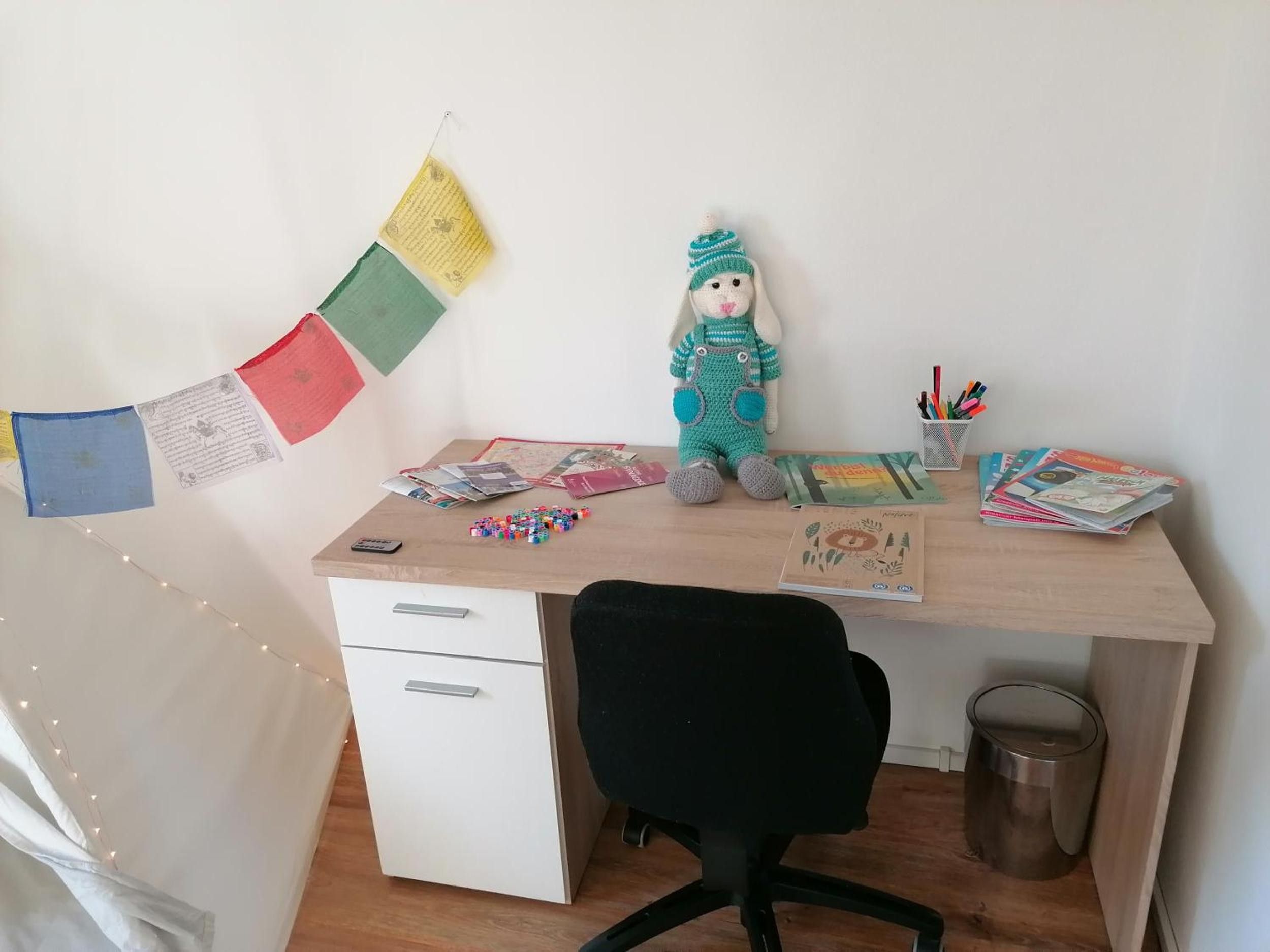 Ein Arbeitsplatz mit Schreibtisch, Stuhl und verschiedenen Gegenständen, darunter ein Stofftier, Papier und ein Mülleimer.