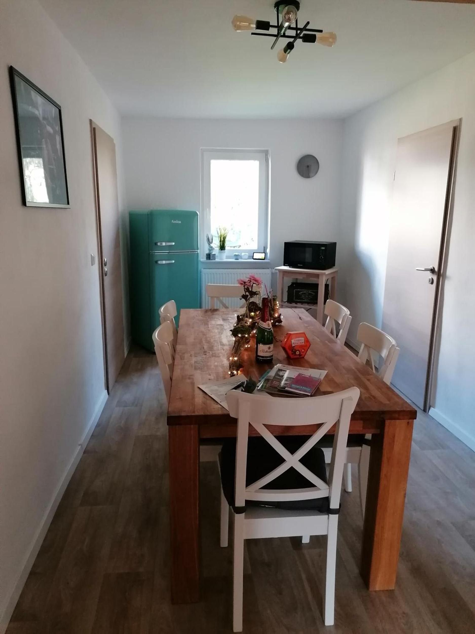 Ein Essbereich mit einem Holztisch, weißen Stühlen, einem türkisblauen Kühlschrank, einem Fernseher auf einem Schrank und einem Fenster mit Tageslicht.