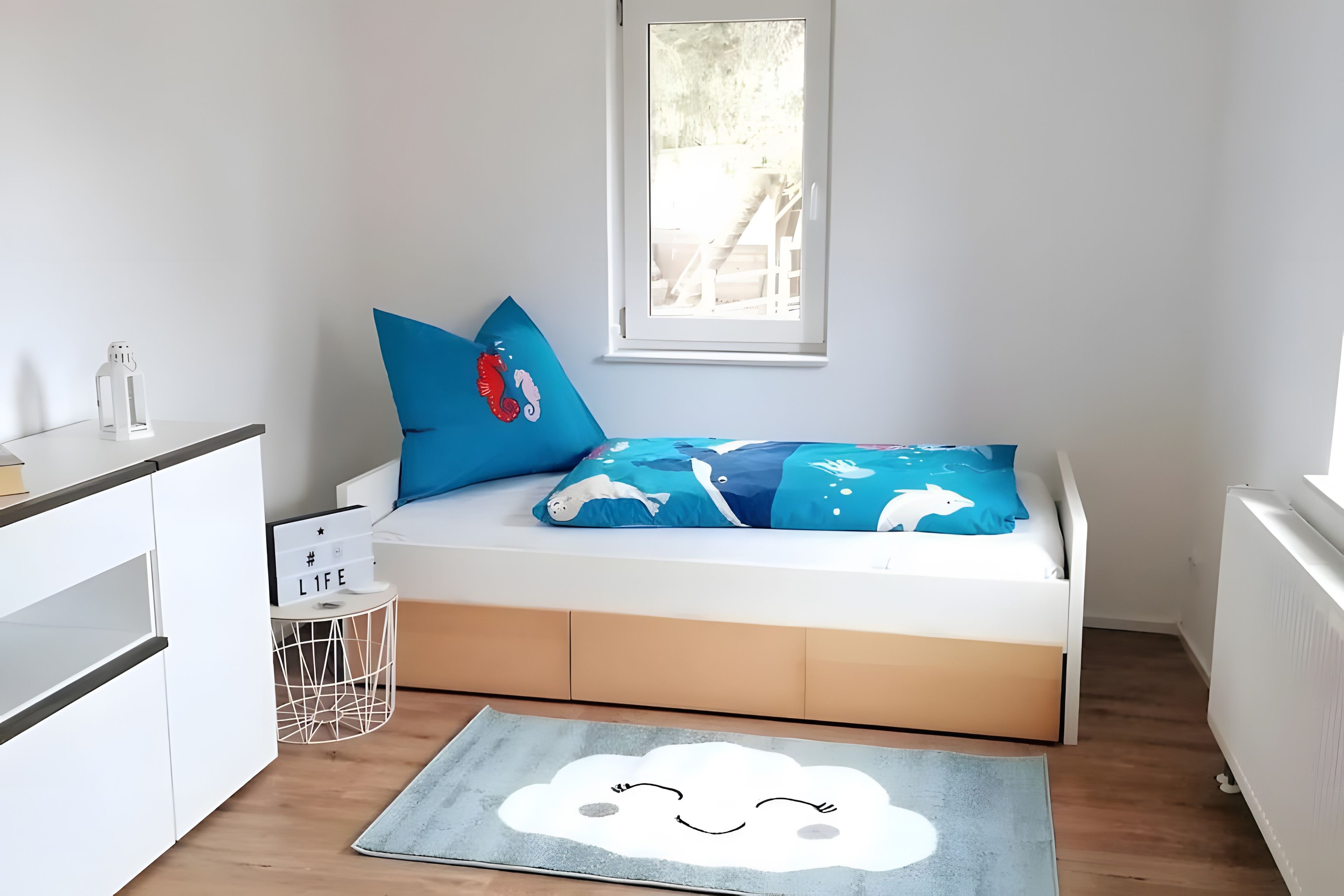 Ein Kinderzimmer mit Bett, Kissen, einem kleinen Nachttisch und einem Fenster.
