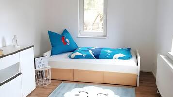 Ein Kinderzimmer mit Bett, Kissen, einem kleinen Nachttisch und einem Fenster.
