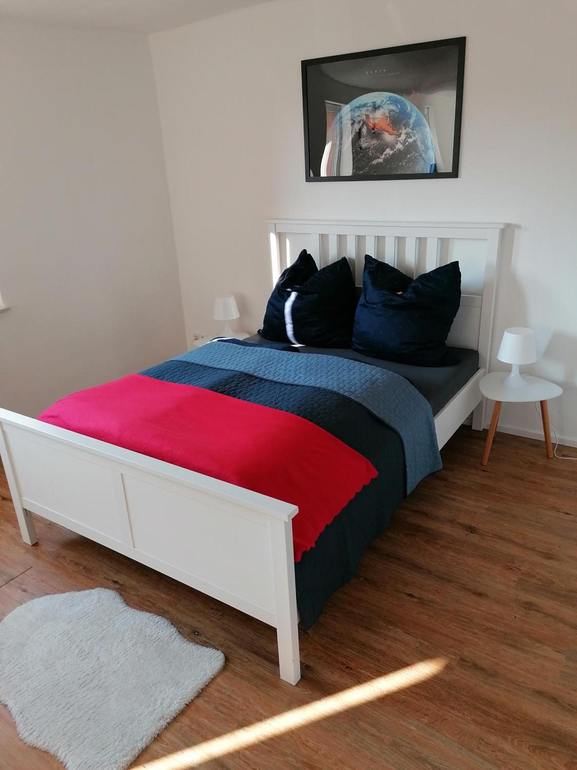 Ein Schlafzimmer mit einem weißen Bett, einer roten und blauen Decke, einem weißen Teppich, einem Nachttisch mit einer Lampe und einem gerahmten Bild an der Wand.
