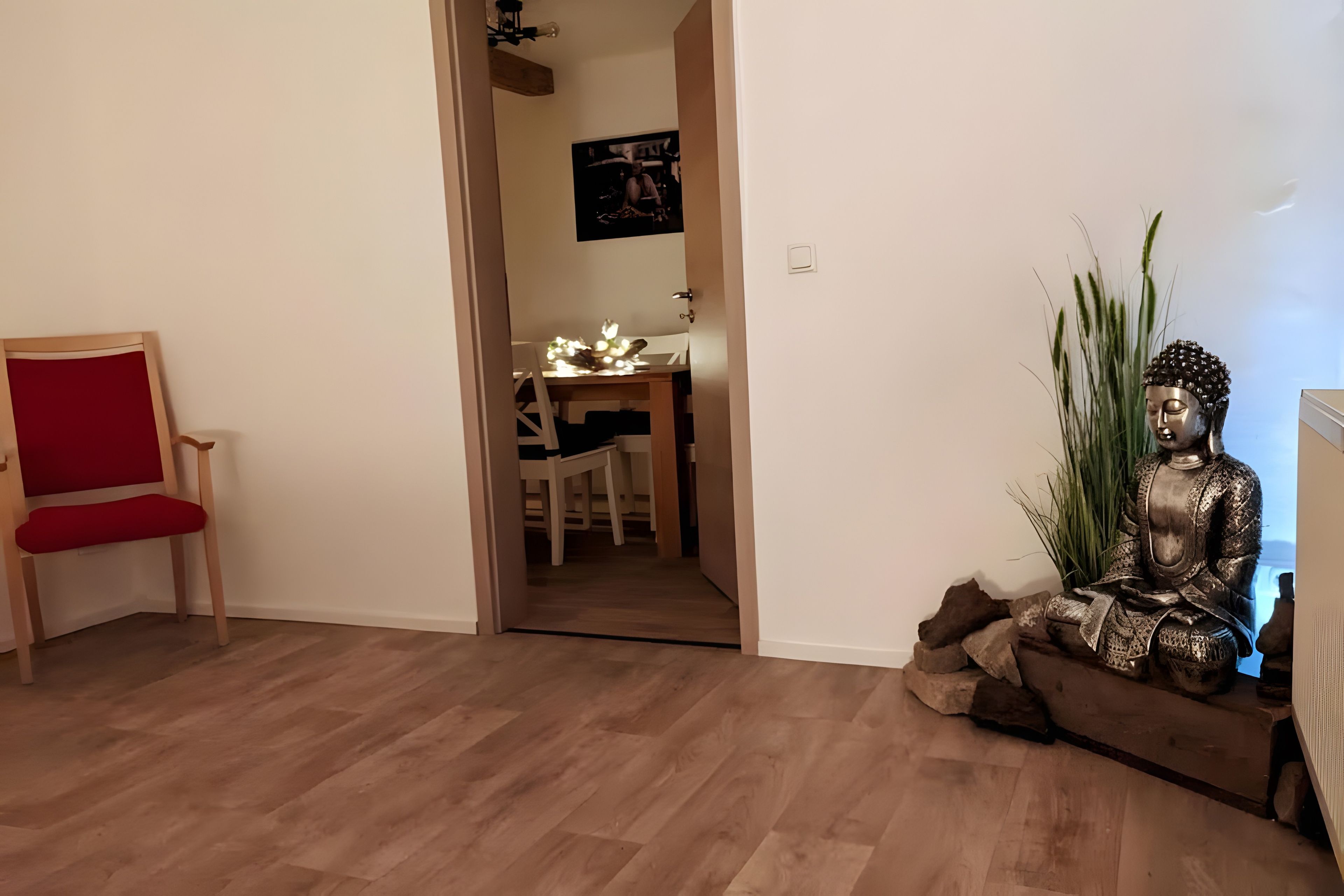 Ein Zimmer mit Holzboden, einem roten Sessel, einer Buddha-Statue und einem Essbereich im Hintergrund.