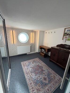 A room with a patterned carpet, a round window, a radiator, a wooden cabinet, and a sewing machine. - New Listing (Schwarzenbach am Wald)