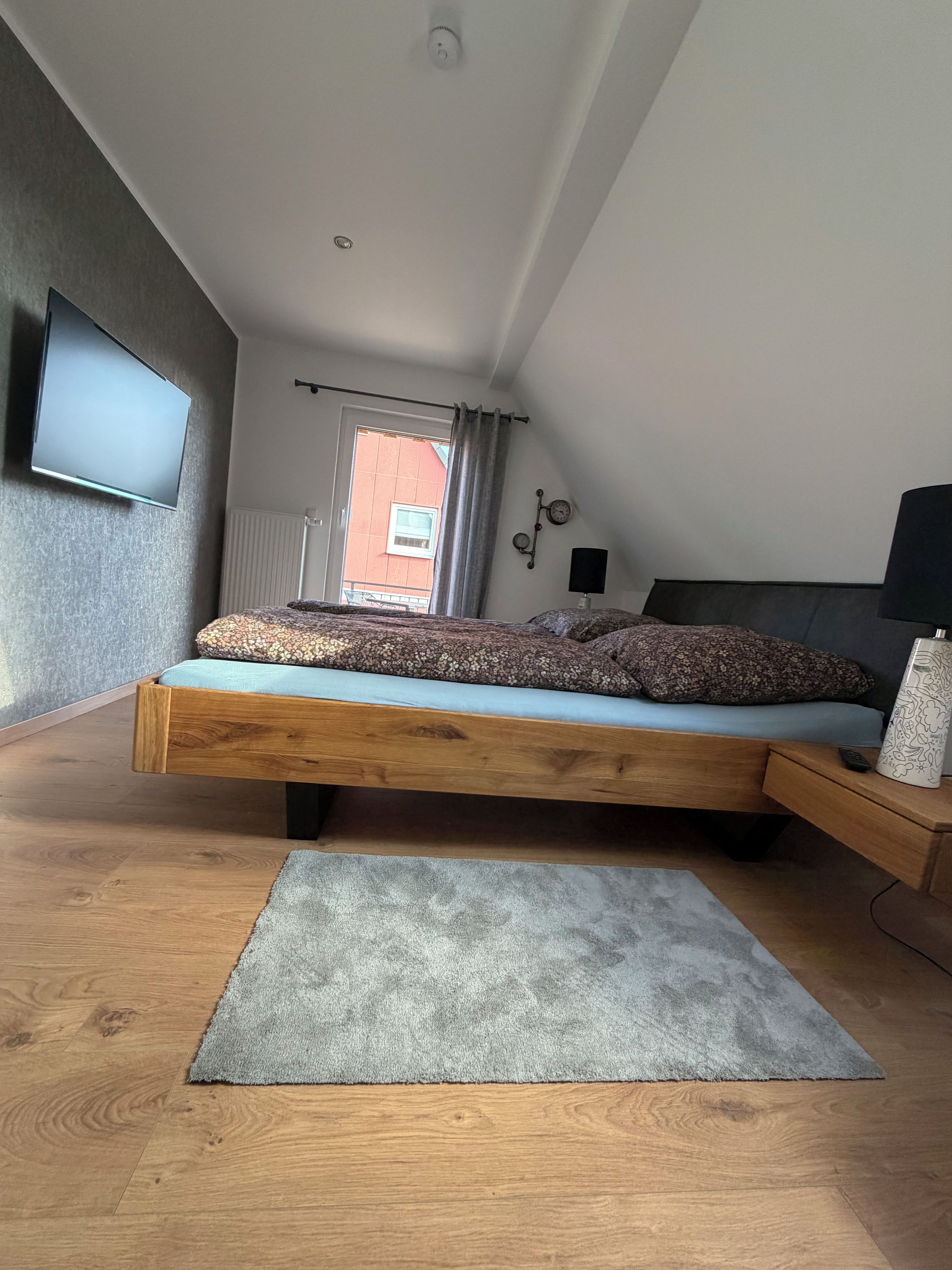 Un dormitorio moderno con cama estilo altillo, un televisor de pantalla plana, una lámpara de noche y vistas a un edificio rojo en el exterior.