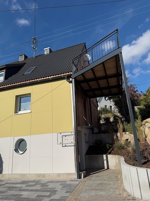 Een gebouw met twee verdiepingen, een geel-witte gevel, een balkon met een metalen hekwerk en een klein raam.