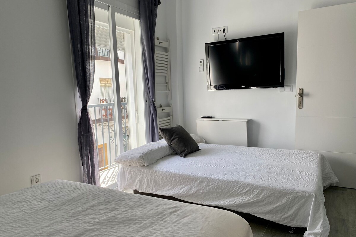 Quarto de hotel com duas camas, uma TV presa à parede e varanda com cortinas.