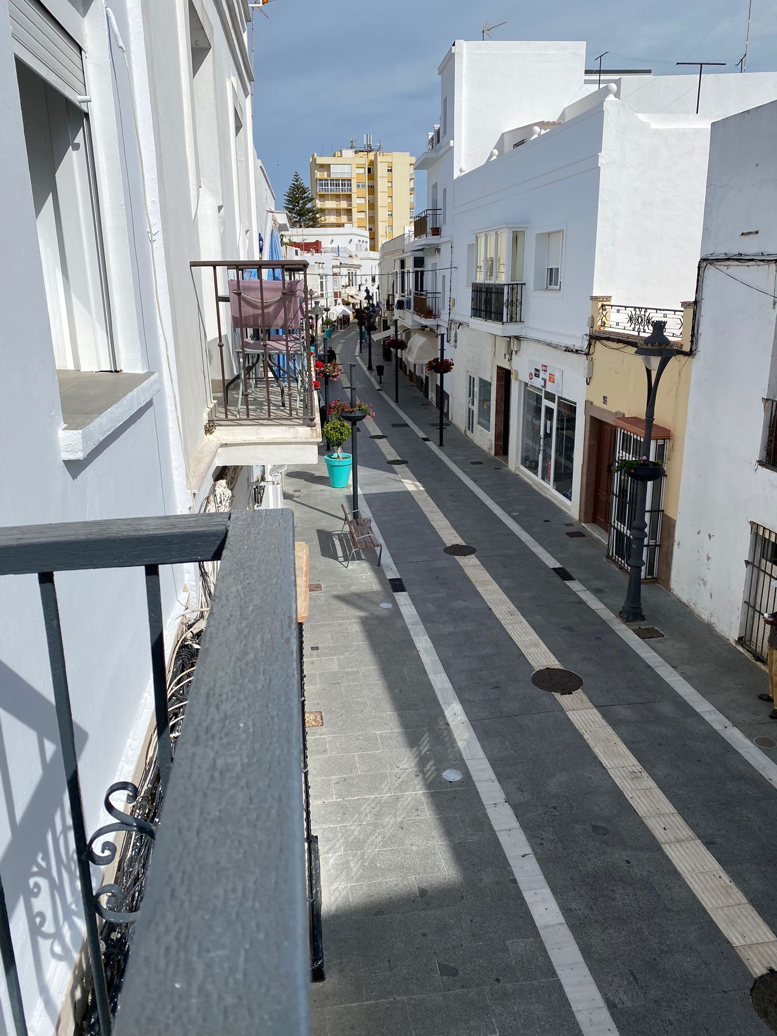 A narrow street with white buildings, a balcony with a metal railing, and a clear sky.